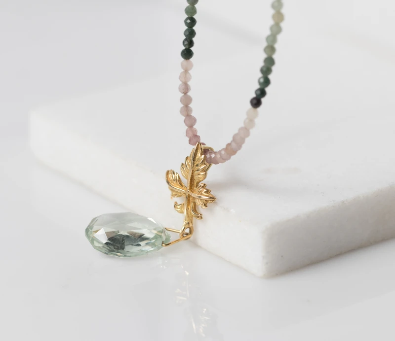 Gold pendant depicting a realistic vine leaf with a shimmering amethyst green pear-shaped stone. The necklace consists of agate stones in pink and green colors and a gold clasp
