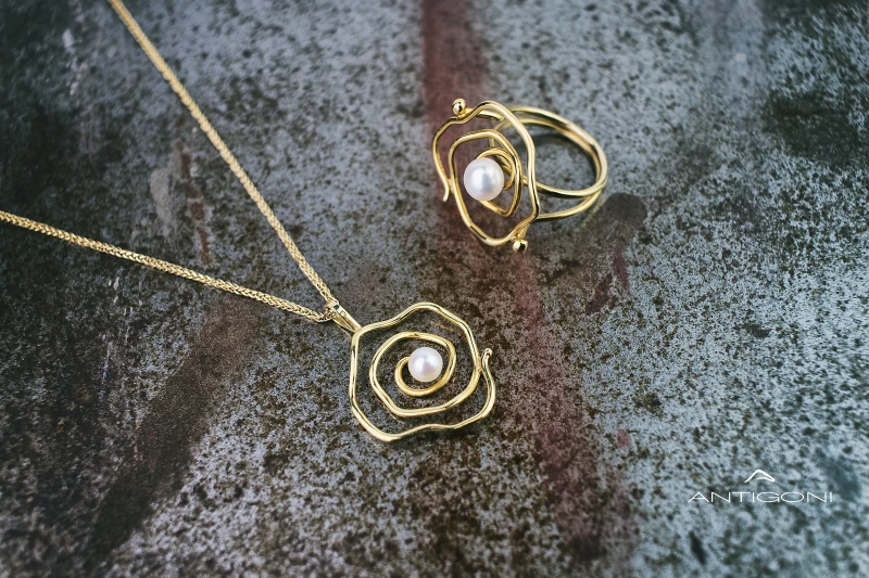 Gold medallion hanging on a fine gold chain. The pendant has a spiral shape and a beautiful round white diamond adorns it in the middle. Next to the pendant is depicted a ring with the same spiral pattern and in the middle is a white natural pearl